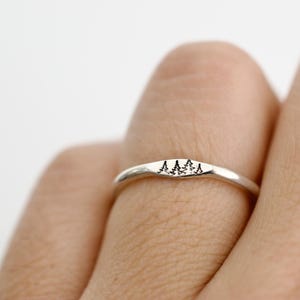 May include: A silver ring with a minimalist design featuring three small trees. The ring is worn on a finger, highlighting its delicate details and nature-inspired theme. The ring's band is smooth and polished, reflecting light.