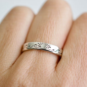 May include: A silver ring with a sun and waves design. The ring is worn on a finger.