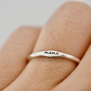 May include: A silver ring engraved with the word "mama" in a cursive script. The ring has a minimalist design with a gently curved band. The ring is displayed on a finger, set against a soft, neutral backdrop.