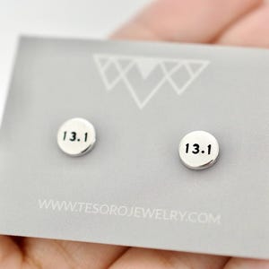 Puede incluir: Pendientes de botón plateados con el número "13.1" en negro. Se muestran en una tarjeta gris con un diseño geométrico y la dirección web www.tesorojewelry.com.