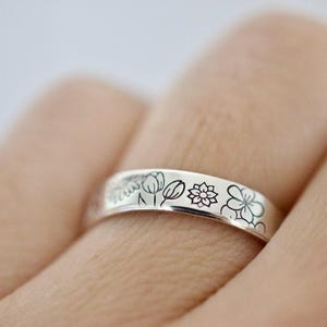 May include: A silver band ring with a floral design engraved around the band. The flowers are outlined in black and include tulips, a clover, and a stylized flower.