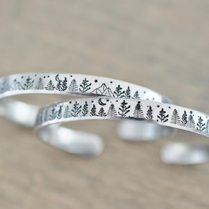 May include: Two silver-colored cuff bracelets. Each bracelet features a detailed forest scene with trees, mountains, stars, and a crescent moon. The bracelets are open-ended and designed to be adjustable.