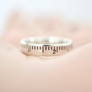 May include: A silver ring with a ruler design engraved on the band. The ruler markings go up to 2 inches.