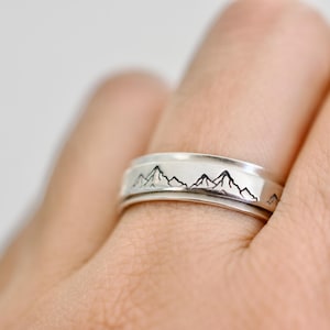 May include: A silver ring with a mountain range design. The ring features a detailed black mountain silhouette around the band. The ring is worn on a finger, showcasing the intricate design and craftsmanship.