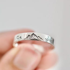 Könnte beinhalten: Ein silberner Ring mit einem Bergketten-, Sonnen- und Wellen-Design. Der Ring wird zwischen den Fingern gehalten und weist eine detaillierte Landschaftsgravur auf. Das Design des Rings umfasst eine Bergkette, eine Sonne und eine Welle.