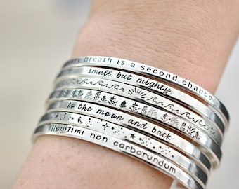 May include: A collection of silver metal bracelets, each engraved with motivational quotes and designs. The bracelets display phrases such as "My breath is a second chance" and "small but mighty". The bracelets are approximately 0.64 cm wide.