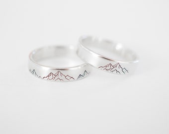 May include: Two silver wedding bands with a mountain range design engraved on each. One band has a red mountain range and the other has a blue mountain range.