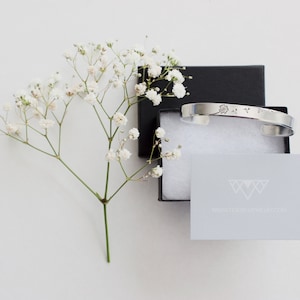 May include: A silver bangle bracelet with a delicate floral design, packaged in a black box with white tissue paper and a small card with the text "www.daintyjewelry.com".