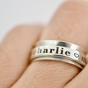 Peut inclure: Une bague en argent avec le nom "charlie" gravé sur le bandeau, ainsi qu'un petit symbole de cœur. La bague est portée au doigt, mettant en valeur son design. La bague est un bijou.