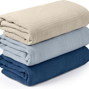K&ouml;nnte beinhalten: Ein Stapel aus drei gefalteten Decken in verschiedenen Farben. Die oberste Decke ist hellbeige, die mittlere ist hellblau und die untere ist dunkelblau. Die Decken scheinen aus einem strukturierten Stoff zu sein.
