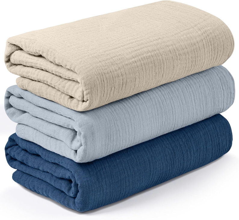 K&ouml;nnte beinhalten: Ein Stapel aus drei gefalteten Decken in verschiedenen Farben. Die oberste Decke ist hellbeige, die mittlere ist hellblau und die untere ist dunkelblau. Die Decken scheinen aus einem strukturierten Stoff zu sein.