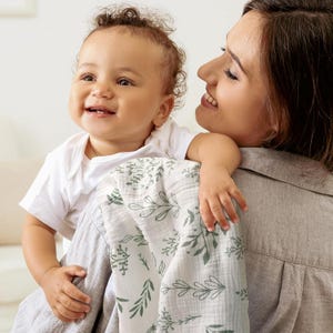 K&ouml;nnte beinhalten: Ein Baby in einem wei&szlig;en Body wird von einer Person gehalten, mit einer wei&szlig;-gr&uuml;nen Decke mit floralem Muster. Die Decke hat ein zartes Design aus Bl&auml;ttern und Zweigen. Das Baby l&auml;chelt.