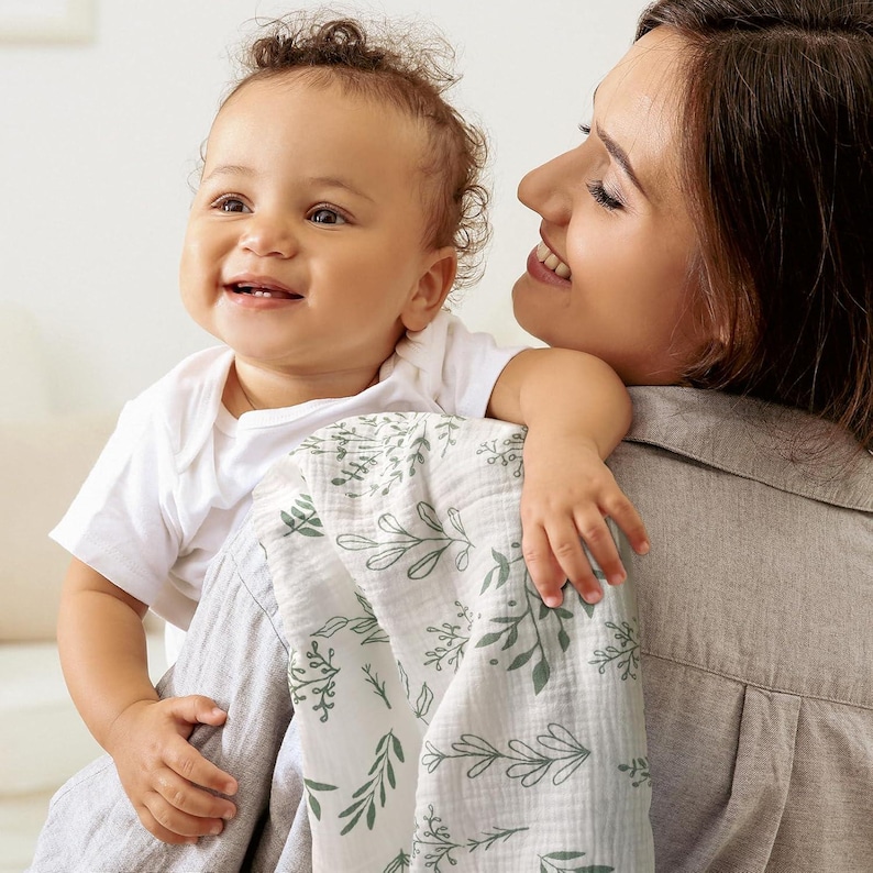 K&ouml;nnte beinhalten: Ein Baby in einem wei&szlig;en Body wird von einer Person gehalten, mit einer wei&szlig;-gr&uuml;nen Decke mit floralem Muster. Die Decke hat ein zartes Design aus Bl&auml;ttern und Zweigen. Das Baby l&auml;chelt.