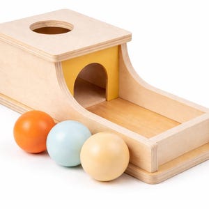May include: Wooden toy with a light wood finish, featuring a ramp and a hole on top. Includes three colorful balls: orange, light blue, and cream. The ramp has a yellow interior.
