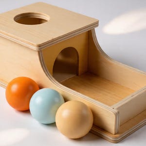 May include: Wooden toy with a box and ramp design, featuring a circular opening on top and a curved opening on the side. Three colourful balls in orange, light blue, and tan are positioned in front of the toy. This educational toy is designed for toddlers.
