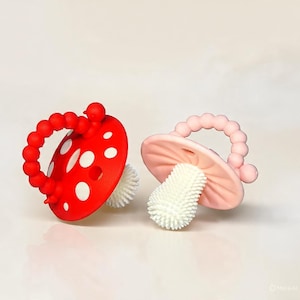 May include: Two mushroom-shaped baby teethers. One teether is red with white spots and a red beaded handle. The other is pink with a pink beaded handle. Both have a white, textured stem.