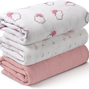 K&ouml;nnte beinhalten: Ein Stapel aus drei gefalteten Decken. Die obere Decke ist wei&szlig; mit einem sich wiederholenden Muster aus rosa Sonnen und Wolken. Die mittlere Decke ist wei&szlig; mit einem Muster aus rosa Kreuzen und grauen Sternen. Die untere Decke ist einfarbig rosa.