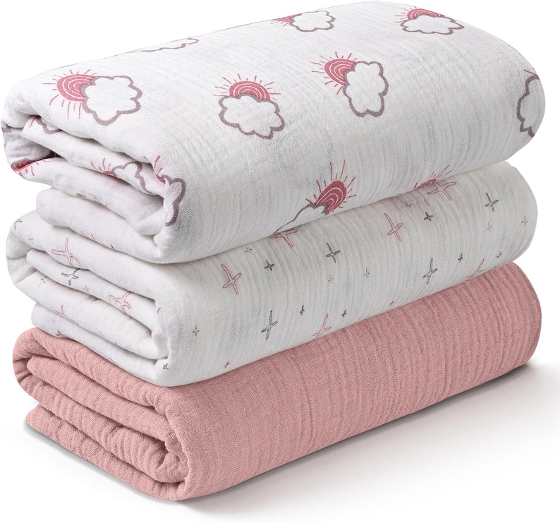 K&ouml;nnte beinhalten: Ein Stapel aus drei gefalteten Decken. Die obere Decke ist wei&szlig; mit einem sich wiederholenden Muster aus rosa Sonnen und Wolken. Die mittlere Decke ist wei&szlig; mit einem Muster aus rosa Kreuzen und grauen Sternen. Die untere Decke ist einfarbig rosa.