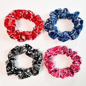 May include: Four scrunchies in red, blue, black, and pink, each with a paisley bandana pattern. The scrunchies are circular and made of fabric, perfect for tying up hair. The red scrunchie is at the top left, the blue at the top right, the black at the bottom left, and the pink at the bottom right.
