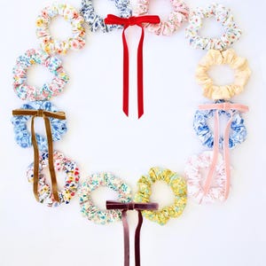May include: A collection of twelve fabric hair scrunchies arranged in a circular wreath shape. The scrunchies feature various floral patterns in pastel and bright colors, with three scrunchies accented by long velvet ribbons in red, brown, and pink.