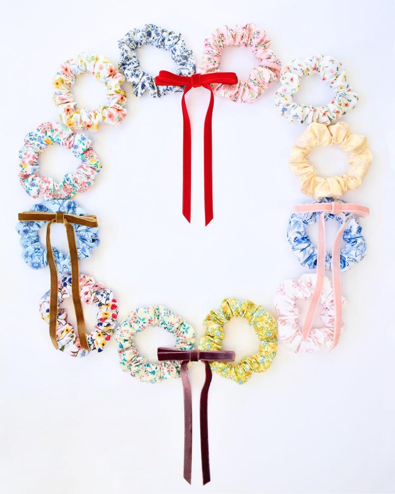 May include: A collection of twelve fabric hair scrunchies arranged in a circular wreath shape. The scrunchies feature various floral patterns in pastel and bright colors, with three scrunchies accented by long velvet ribbons in red, brown, and pink.