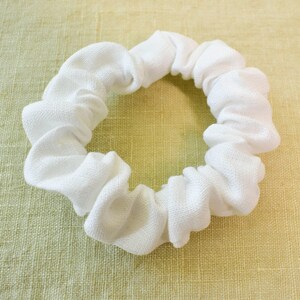 May include: A white fabric scrunchie for hair. The scrunchie is made of a soft, textured fabric and is designed to be gentle on hair.