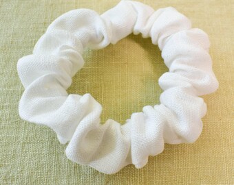 May include: A white fabric scrunchie for hair. The scrunchie is made of a soft, textured fabric and is designed to be gentle on hair.