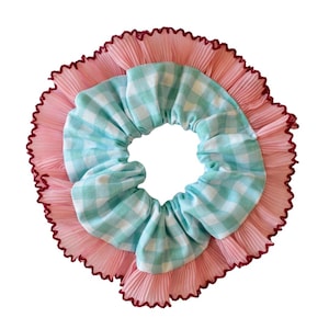 May include: A scrunchie with a light blue and white gingham pattern center, surrounded by a pink pleated fabric. The outer edge is trimmed with a dark red, ruffled detail. This hair accessory is designed for everyday wear.