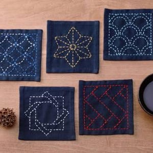 May include: Six square blue fabric coasters with different embroidered designs in white, yellow, and blue thread. The coasters are on a wooden surface.