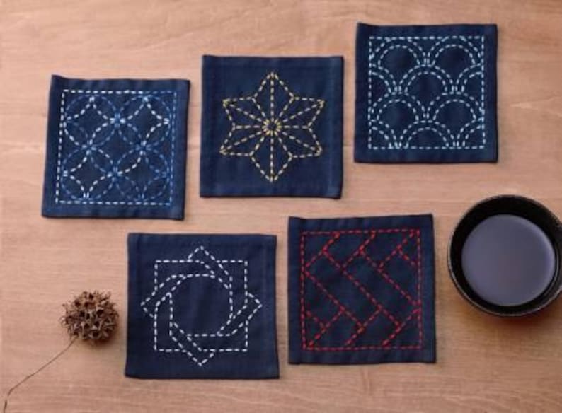 May include: Six square blue fabric coasters with different embroidered designs in white, yellow, and blue thread. The coasters are on a wooden surface.