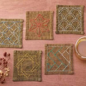 May include: Five brown fabric coasters with different embroidered designs. The designs include a geometric pattern, a leaf, a wave pattern, a zig-zag pattern, and a square pattern.