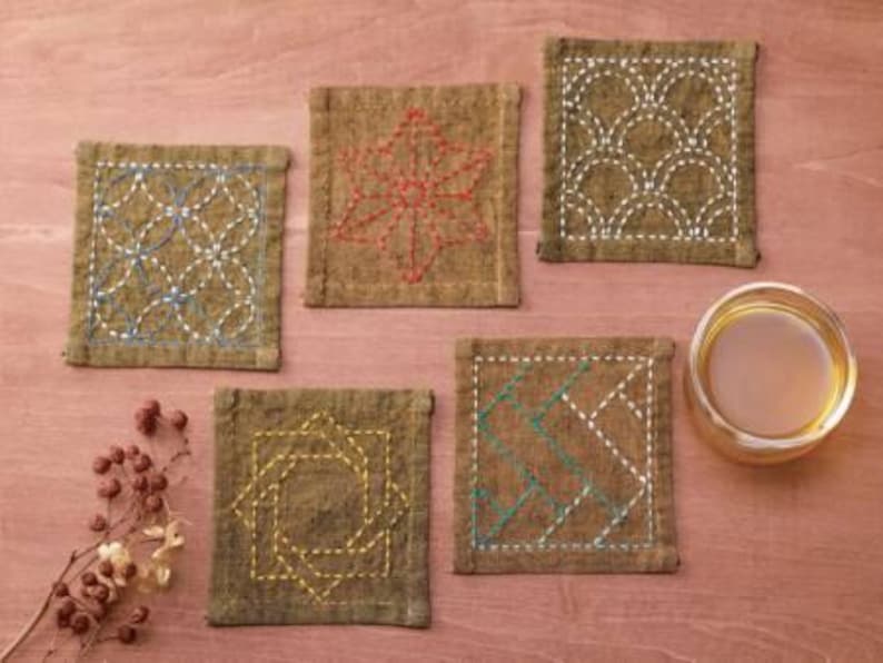 May include: Five brown fabric coasters with different embroidered designs. The designs include a geometric pattern, a leaf, a wave pattern, a zig-zag pattern, and a square pattern.
