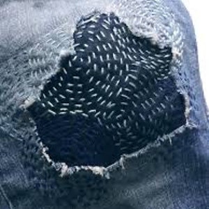 K&ouml;nnte beinhalten: Nahaufnahme einer blauen Jeanshose mit einem gro&szlig;en, dunkelblauen Flicken, der &uuml;ber ein Loch am Knie gen&auml;ht ist. Der Flicken ist mit wei&szlig;en N&auml;hten in einem wirbelnden Muster verziert.