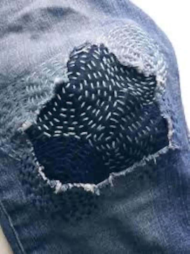 K&ouml;nnte beinhalten: Nahaufnahme einer blauen Jeanshose mit einem gro&szlig;en, dunkelblauen Flicken, der &uuml;ber ein Loch am Knie gen&auml;ht ist. Der Flicken ist mit wei&szlig;en N&auml;hten in einem wirbelnden Muster verziert.
