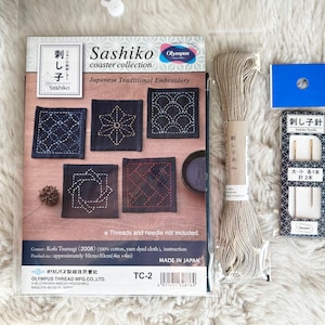May include: A Sashiko coaster collection kit with five pre-stitched coasters, embroidery floss, two needles, and instructions. The coasters are made of dark blue fabric with white stitching in geometric patterns. The kit is labeled "Sashiko Coaster Collection" and "Japanese Traditional Embroidery".