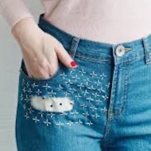 K&ouml;nnte beinhalten: Blaue Jeans aus Denim mit einem wei&szlig;en Patch auf der vorderen Tasche, verziert mit wei&szlig;en Kreuzstich-Stickereien.