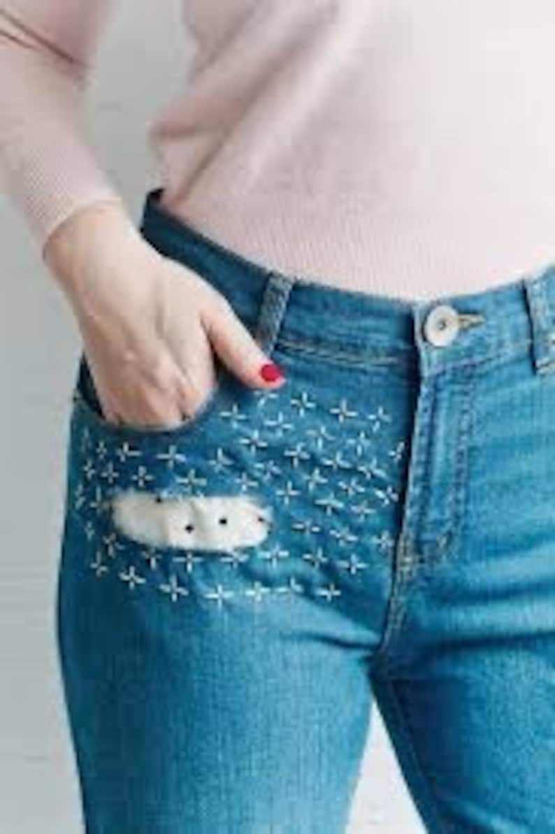 K&ouml;nnte beinhalten: Blaue Jeans aus Denim mit einem wei&szlig;en Patch auf der vorderen Tasche, verziert mit wei&szlig;en Kreuzstich-Stickereien.