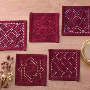 May include: Six square-shaped coasters with different embroidered designs. The coasters are made of a dark red fabric with white stitching. The designs include a star, a geometric pattern, a wave pattern, and a diamond pattern.