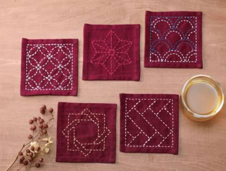 May include: Six square-shaped coasters with different embroidered designs. The coasters are made of a dark red fabric with white stitching. The designs include a star, a geometric pattern, a wave pattern, and a diamond pattern.