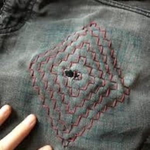 K&ouml;nnte beinhalten: Nahaufnahme einer Jeansjacke mit einem rautenf&ouml;rmigen Patch aus roten N&auml;hten. Die N&auml;hte sind in einem Gittermuster angeordnet und haben ein kleines Loch in der Mitte.