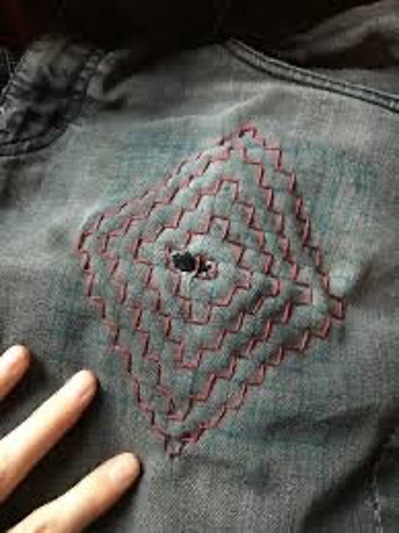 K&ouml;nnte beinhalten: Nahaufnahme einer Jeansjacke mit einem rautenf&ouml;rmigen Patch aus roten N&auml;hten. Die N&auml;hte sind in einem Gittermuster angeordnet und haben ein kleines Loch in der Mitte.