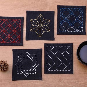 May include: Five black fabric coasters with different embroidered designs. The designs include a starburst, a geometric pattern, a diamond pattern, a circle with a square inside, and a wave pattern. The coasters are on a wooden surface.