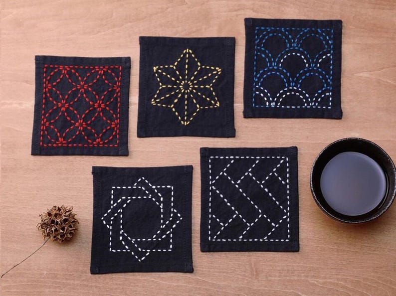 May include: Five black fabric coasters with different embroidered designs. The designs include a starburst, a geometric pattern, a diamond pattern, a circle with a square inside, and a wave pattern. The coasters are on a wooden surface.