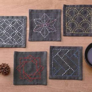 May include: Six gray fabric coasters with different embroidered designs in white, yellow, and red thread. The coasters are on a wooden surface.