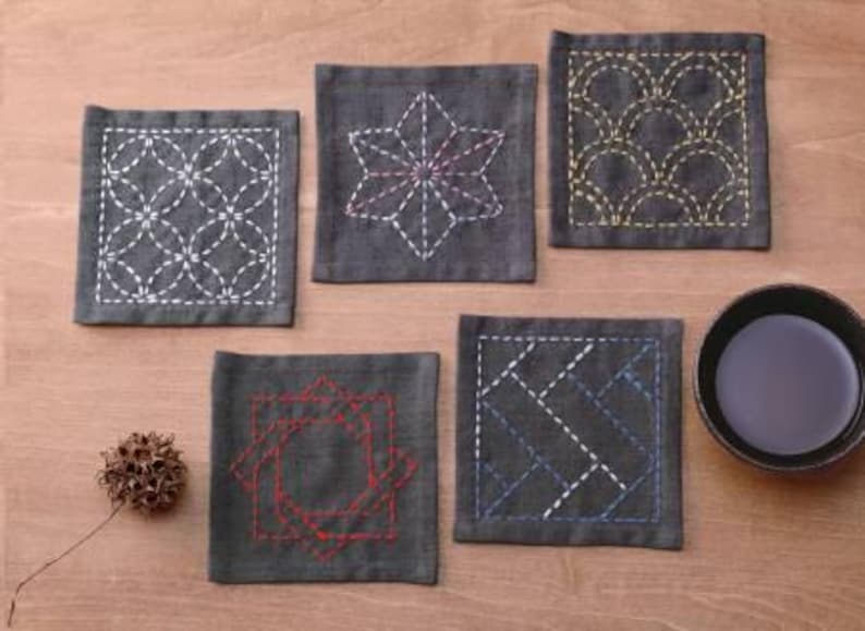 May include: Six gray fabric coasters with different embroidered designs in white, yellow, and red thread. The coasters are on a wooden surface.