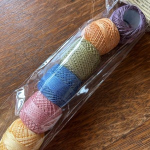 May include: A package of colorful embroidery thread spools in shades of yellow, pink, blue, green, orange, and purple. The spools are in a clear plastic sleeve. The packaging includes the text "AM A COLONIAL".