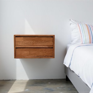 May include: A floating, two-drawer wooden nightstand with a natural finish. The nightstand is mounted on a white wall, next to a bed with white bedding and a striped pillow. The wood grain is visible on the drawers.