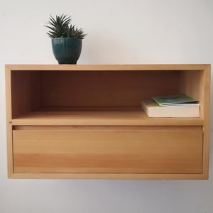 Può includere: Un ripiano a muro in legno chiaro con un cassetto e uno scaffale aperto. Una piccola pianta verde in vaso si trova sullo scaffale superiore. Un libro è sullo scaffale aperto.