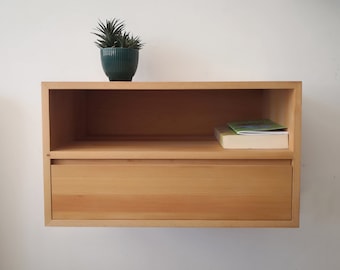May include: A light wood wall mounted shelf with a drawer and an open shelf. A small green potted plant sits on the top shelf. A book is on the open shelf.