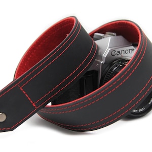 May include: Black leather camera strap with red stitching. The strap is wrapped around a silver Canon camera.